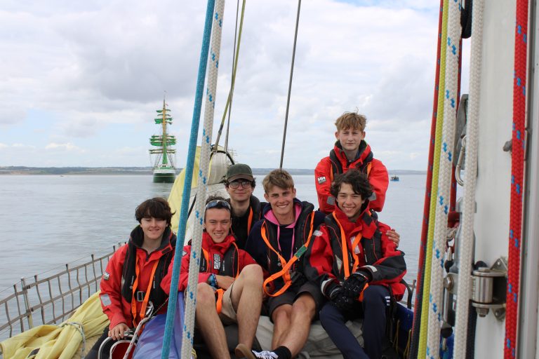 Nathan and fellow crew mates pose on Alba Explorer for a photograph. In the background, you can see a 3-mast tall ship.