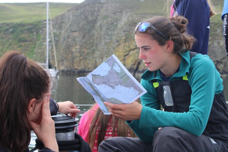 New cadet, Hannah Staal, sits aboard Alba Explorer with a print-out of a map. In the background, there is a sheer cliff face and other vessels.