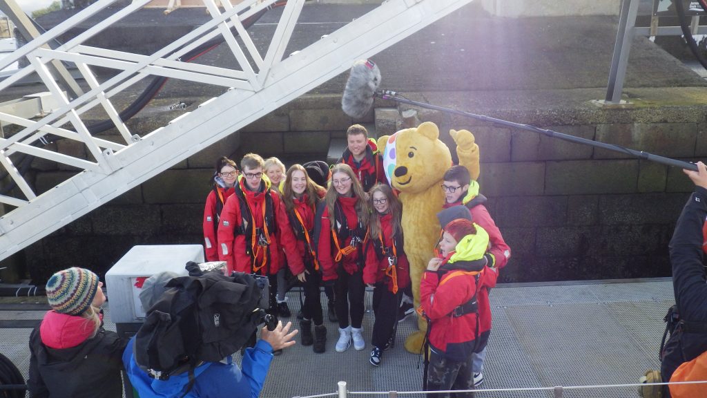 Group of young carers stand on the pontoon of James Watt Dock with Pudsey the bear. There is a film crew in front of them taking a picture.