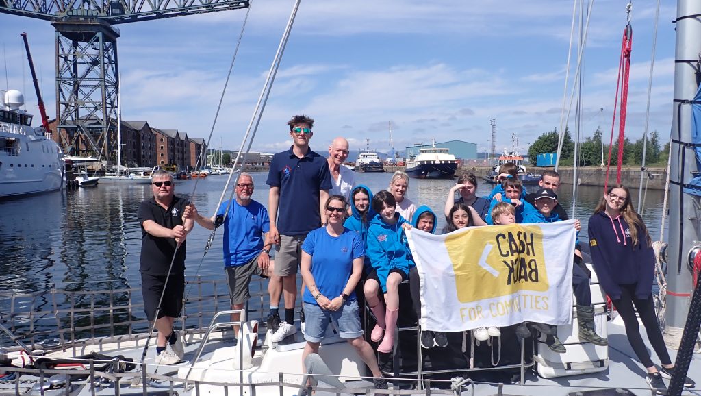 Young people stand aboard Alba Venturer holding a white flag with the yellow 'Cashback for Communities' logo.