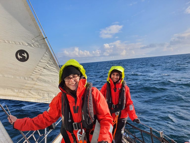 Two young people pose at the front of the boat in their red and green waterproofs. In the background, it is clear skies and choppy waters.
