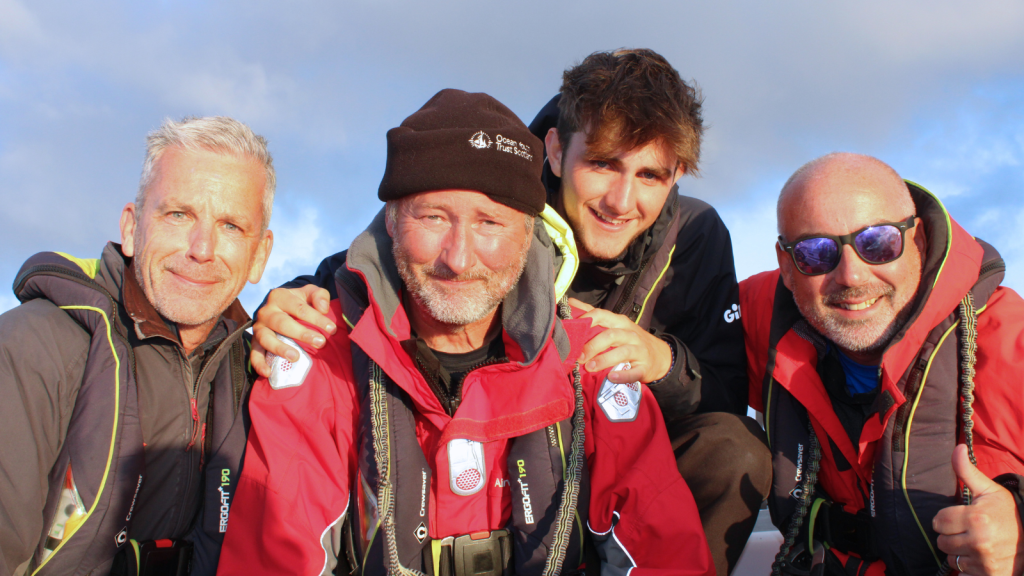 Trustee. Paul, sits alongside fellow volunteers, Iain and Pablo on a boat. First Mate, Wil, is smiling over Paul's shoulder. They are all wearing waterproofs and lifejackets.