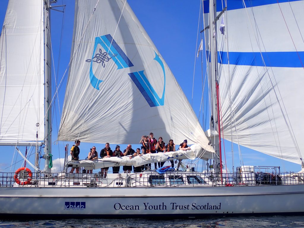 Picture of Alba Venturer with her sails up on a clear, calm day. The crew are standing at the boom and smiling to the camera (which is on another boat).